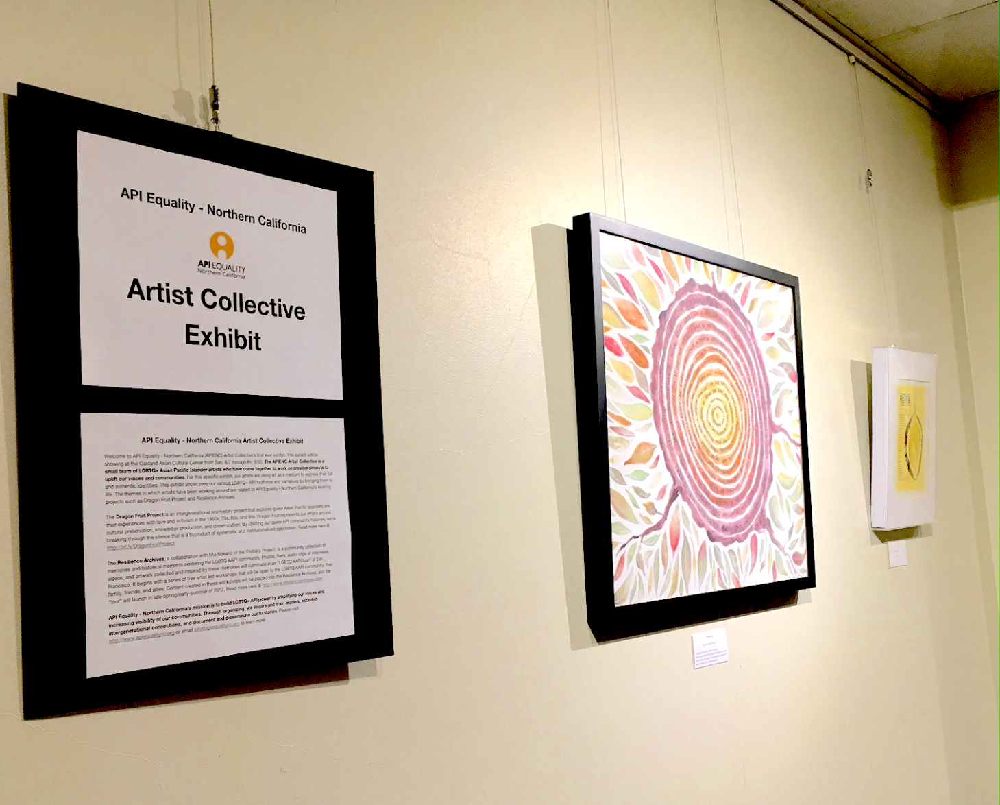 Image Description: The opening section of the Artist Collective exhibit shows three pieces hanging. A statement from APIENC is seen on the left, with a colorful piece in the middle, and a smaller yellow print on the right.
