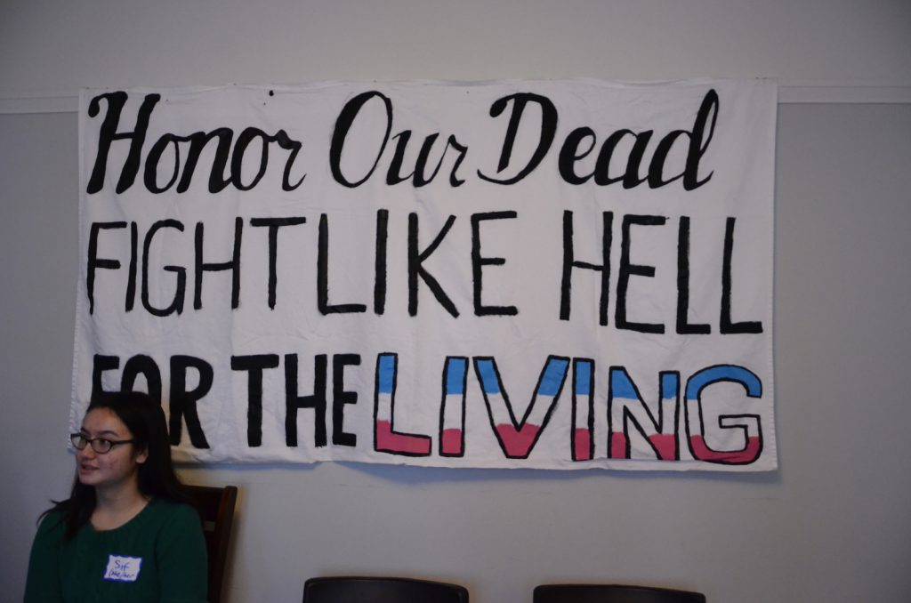 [Image description: A volunteer is sitting to the left of a banner reading, "Honor Our Dead. Fight Like Hell for the Living."
