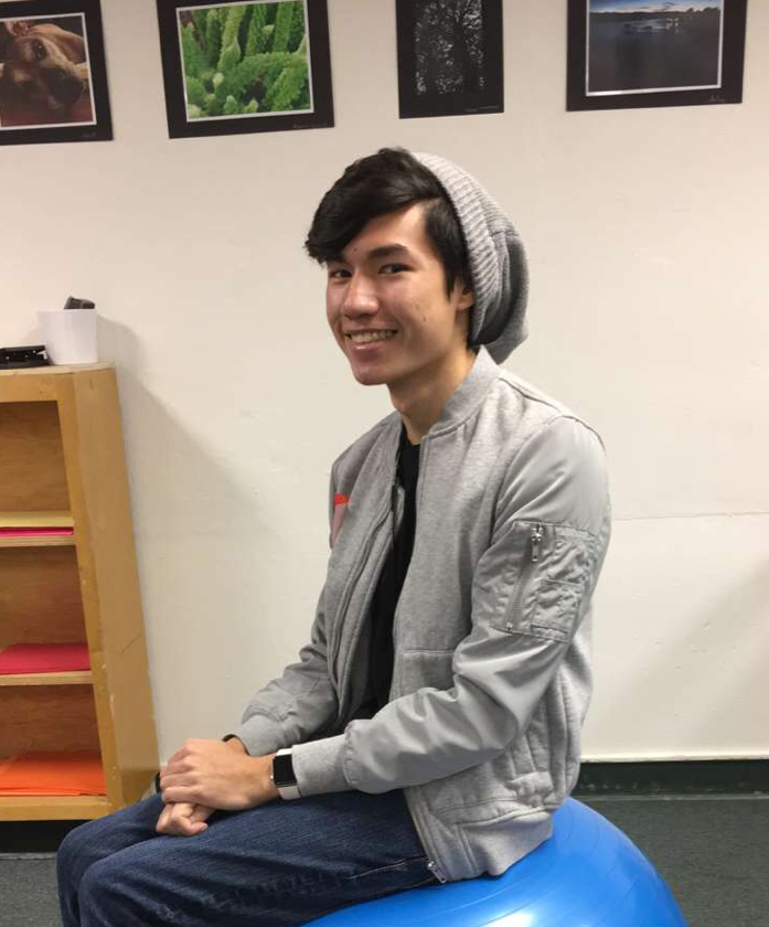 Image Description: Calvin, wearing a grey beanie and grey jacket, smiles while sitting on a blue exercise ball.