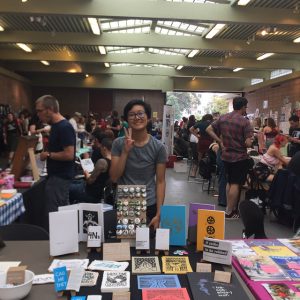 Yi-Yi stands behind a table, smiling, and displaying peace sign with their right hand. On the table are zines and artwork on display.