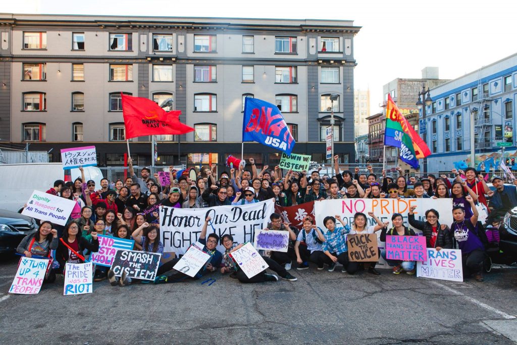 Image Description: A large group of LGBTQ API people and allies pose with banners, flags, and signs at the 2016 Trans March. They have raised fists, and are looking fiercely at the camera.