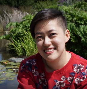 Image description: Nancy is outside, smiling toward the camera. She is wearing a red floral shirt. In back of her is a pond with plantlife growing in it.
